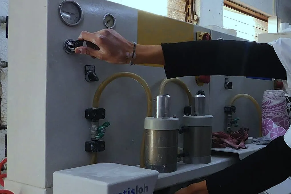 Mano girando una perilla en el panel de una máquina Satisloh, con manómetros, mangueras amarillas y dos cilindros