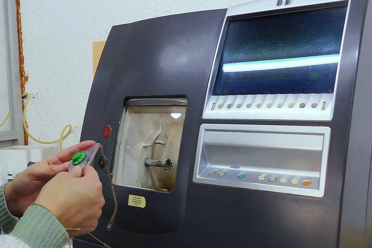 Manos acercando un armazón con su lente a una biseladora automática, con ventana de trabajo y panel de controles